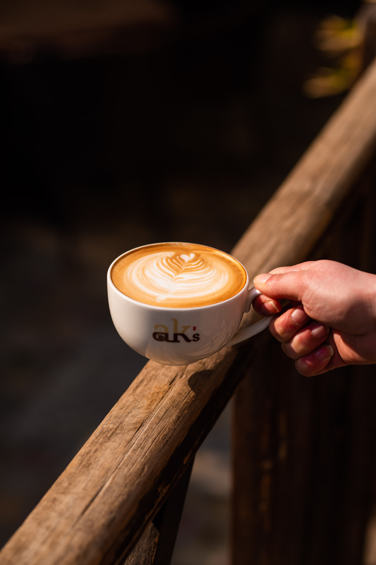 A hand holding an AK's cappuccino on a wooden rail